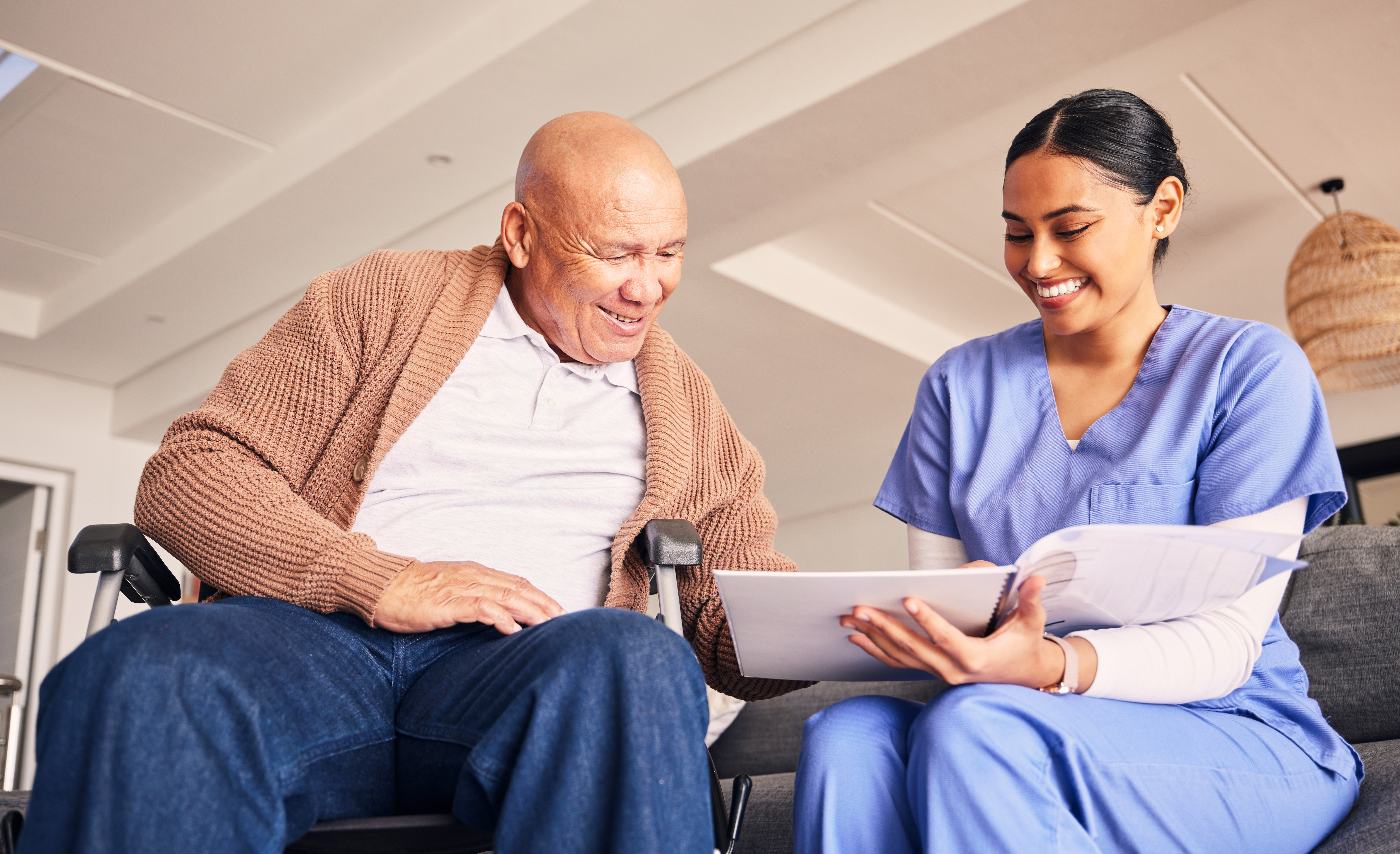 A smiling elderly man in a wheelchair sits next to a young female healthcare worker in scrubs. They are looking at a document together and appear to be having a pleasant conversation in a bright, modern living space.