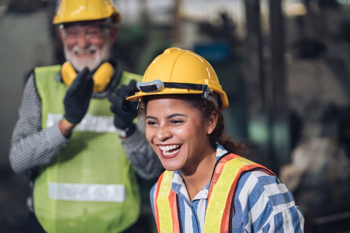 Two construction workers, a man and a woman, wearing hard hats and safety vests, working on a construction site. The woman is smiling and laughing, while the man has a friendly, approving expression on his face. The woman is wearing a striped shirt and the man is wearing a checkered shirt. Industrial equipment and structures are visible in the background.